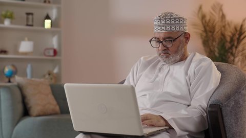 تصفح الانترنت وبرامج التواصل الاجتماعي، تطور التكنولوجيا والتقنيات الحديثة، استخدام الحاسوب المحمول لإنجاز المهام، بورتريه لرجل كبير السن عربي خليجي عماني يرتدي الدشداشة و الكمة و اناظارة الطبية يجلس على الكرسي يحمل بيده جهاز اللابتوب