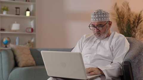 إجراء مكالمات الفيديو، تطور التكنولوجيا والتقنيات الحديثة، استخدام الحاسوب المحمول لإنجاز المهام، بورتريه لرجل كبير السن عربي خليجي عماني يرتدي الدشداشة و الكمة و النظاره الطبية يجلس على الكرسي يحمل بيده جهاز اللابتوب