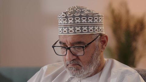 مفهوم توضيح الصورة، ايماءات التركيز والجدية ، تصويرمقرب  لرجل مسن عربي خليجي عماني يرتدي الدشداشة والكمة  و النظارة الطبية يحاول قراءة شي ما ، الفحص والإستكشاف والقراءة