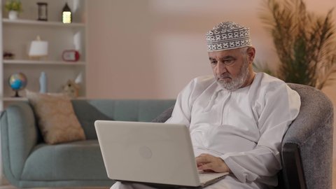 تطور التكنولوجيا والتقنيات الحديثة، استخدام الحاسوب المحمول لإنجاز المهام، بورتريه لرجل كبير السن عربي خليجي عماني يرتدي الدشداشة و الكمة يجلس على الكرسي يحمل بيده جهاز اللابتوب، إجراء مكالمات الفيديو
