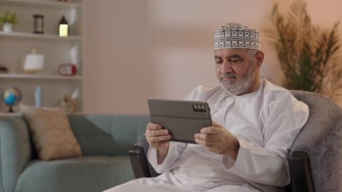 ايماءات التركيز والجدية ، استخدام التكنولوجيا و التقنيات الحديثة, تطبيقات وبرامج الأجهزة اللوحية، مفهوم إدارة المشاريع وانجاز المهام عن بعد، تصوير لرجل مسن عربي خليجي عماني يرتدي الدشداشة والكمة  يجلس على كرسي يحمل جهاز التابلت ، تصفح مواقع و وسائل التواصل الاجتماعي .