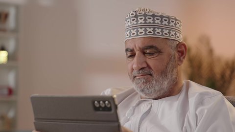 Tablet applications and programs, gestures of focus and seriousness, the use of technology and modern techniques, the concept of project management and completing tasks remotely, a close-up shot of an elderly Arab Gulf Omani man wearing a dishdasha and kumma holding a tablet device, browsing social media sites.