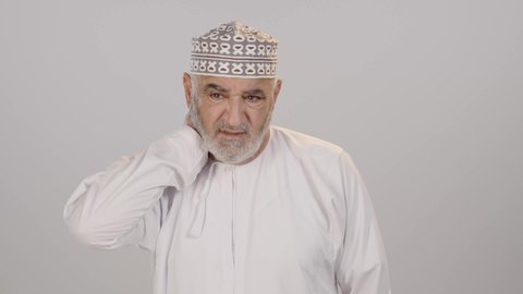 إيماءات الوجه تدل على الاضطراب والتألم، آلام الرقبة والعظام والعمود الفقري، ضرورة الذهاب للطبيب والحصول على العلاج، بورتريه مقرب لرجل مسن عربي خليجي عماني يرتدي الدشداشة و الكمه يمسك رقبته بتعابير الألم، التهاب العضلات والمفاصل، خلفية بيضاء