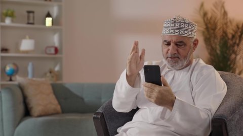 التواصل مع الأهل والأصدقاء، إجراء مكالمات الفيديو عبر الهاتف المحمول، استخدام التكنولوجيا و التقنيات الحديثة, تصويرلرجل مسن عربي خليجي عماني يرتدي الدشداشة و الكمة يجلس على الكرسي يمسك الجوال بايماءات السعاده و السرور