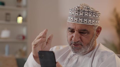 Making video calls via mobile phone, communicating with family and friends, using modern technology and techniques, a close-up shot of an elderly Arab Gulf Omani man wearing a dishdasha and kumma sitting on a chair holding a mobile phone with gestures of happiness and joy.