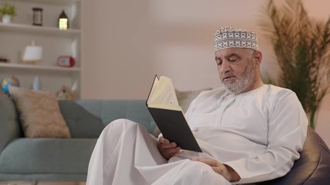 قضاء أوقات الفراغ بالتثقيف والتعلم، حب المطالعة والقراءة، مفهوم التأمل والتفكير, تنمية مهارات القراءة, الاهتمام بتصفح الكتب المفيدة, رجل مسن عربي خليجي عماني يرتدي الدشداشة والكمة يجلس على الاريكة ويمسك بيده الكتاب