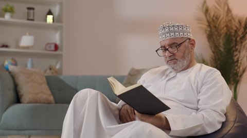 The love of reading and literature, the concept of contemplation and thinking, spending free time on education and learning, developing reading skills, the interest in browsing useful books, an elderly Arab Gulf Omani man wearing a dishdasha and a kumma and reading glasses sitting on the sofa holding a book in his hand.