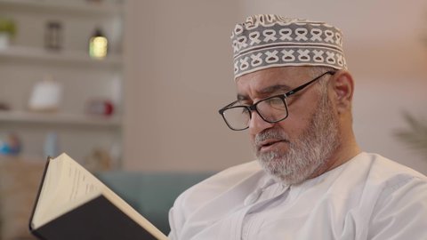 The concept of contemplation and thinking, the love of reading and literature, spending free time on education and learning, developing reading skills, the interest in browsing useful books, a close-up image of an elderly Arab Gulf Omani man wearing a dishdasha and kumma and holding a book in his hand.