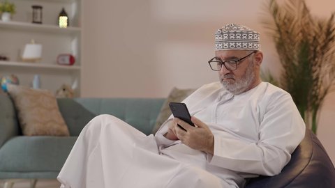 الدردشة مع الأصدقاء عبر برامج وتطبيقات الجوال، استخدام جهاز تقني حديث ومتطور، ايماءات التركيز والجدية، تصوير لرجل مسن عربي خليجي عماني يرتدي الدشداشة والكمة و النظارة الطبية يجلس على الكرسي يحاول قراءة شي ما عبر الجوال