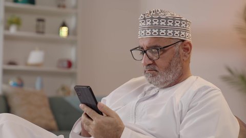 التصفح عبر وسائل التواصل الاجتماعي، الدردشة مع الأصدقاء عبر برامج وتطبيقات الجوال، استخدام جهاز تقني حديث ومتطور، ايماءات التركيز والجدية، تصويرمقرب لرجل مسن عربي خليجي عماني يرتدي الدشداشة والكمة و النظارة الطبية يجلس على الكرسي يحاول قراءة شي ما عبر الجوال