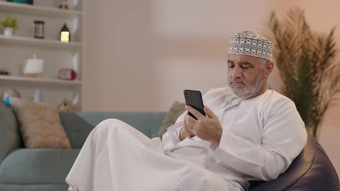 التواصل والدردشة مع الأهل والأصدقاء عبر تطبيقات الجوال، تصفح مواقع التواصل الاجتماعي, رجل مسن عربي خليجي عماني يرتدي الكمة و الدشداشة يجلس على كرسي مريح يقوم باستخدام الهاتف المحمول, استخدام التقنيات الحديثة المتطورة