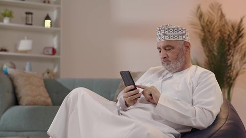 تصفح مواقع التواصل الاجتماعي, التواصل والدردشة مع الأهل والأصدقاء عبر تطبيقات الجوال, رجل مسن عربي خليجي عماني يرتدي الكمة و الدشداشة يجلس على كرسي مريح يقوم باستخدام الهاتف المحمول, استخدام التقنيات الحديثة المتطورة