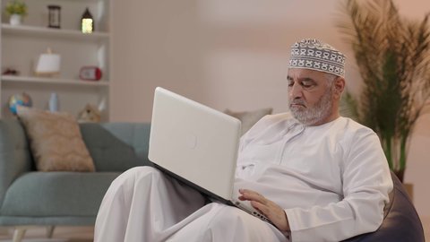 استخدام الحاسوب المحمول لإنجاز المهام، بورتريه لرجل كبير السن عربي خليجي عماني يرتدي الدشداشة و الكمة يجلس على الكرسي يحمل بيده جهاز اللابتوب، تطور التكنولوجيا والتقنيات الحديثة، إجراء مكالمات الفيديو