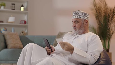 The ease of purchasing and paying through mobile phones, a depiction of an elderly Arab Gulf Omani man wearing a dishdasha and a kumma sitting on the sofa holding a mobile phone and a credit card, paying bills with electronic cards.