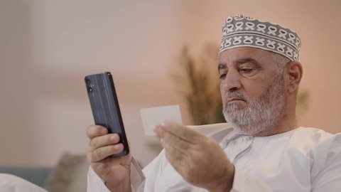 Paying bills with electronic cards, ease of purchasing and payment through mobile phones, a close-up shot of an elderly Arab Gulf Omani man wearing a dishdasha and kumma holding a mobile phone and a credit card in his hand.