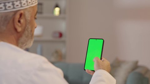 Browsing social media, the development of technology and modern techniques, a close-up portrait from the back of an Arab Gulf Omani man wearing a dishdasha and kumma, displaying a mobile phone with a blank green screen, mobile programs and applications, white background.