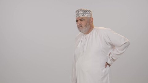 الإحساس بالألم والإصابة بمرض الديسك، بورتريه لرجل مسن عربي خليجي عماني يرتدي الدشداشة و الكمه يقف بصعوبة ويمسك ظهره بيديه بإيماءات المعاناة والوجع، الحاجة للذهاب عند الطبيب لتلقي العلاج، آلام الظهر والعمود الفقري والتهاب المفاصل، خلفية بيضاء