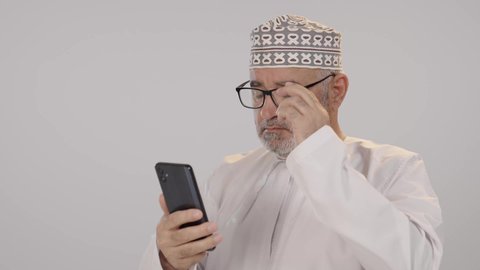 Browse modern communication programs, communicate and chat with family and friends through mobile applications, a close-up portrait of an elderly Arab Gulf Omani man wearing a dishdasha and kumma, wearing glasses, with a white background.