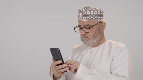 التواصل والدردشة مع الأهل والأصدقاء عبر تطبيقات الجوال، تصفح مواقع التواصل الاجتماعي, بورتريه مقرب لرجل مسن عربي خليجي عماني يرتدي الدشداشة و الكمة يرتدي النظارة الطبية، خلفية بيضاء