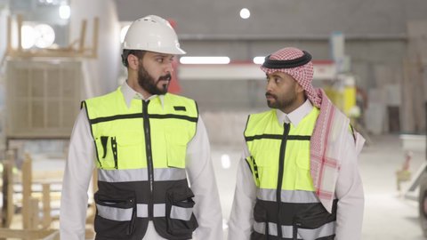 متابعة الأعمال والنقاش حول الأداء، ارتداء خوذة الحمايه في المصنع، تصويرلمهندسان عربيان سعوديان خليجيان يرتديان الثوب يقومان بالمشي في احدى المستودعات ، معامل ومصانع  محليه سعوديه