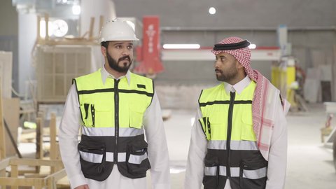 الإشراف على المهام وتبادل الآراء المهنية، ارتداء خوذة الحمايه في المصنع، تصويرلمهندسان عربيان سعوديان خليجيان يرتديان الثوب يقومان بالمشي في احدى المستودعات ، معامل ومصانع  محليه سعوديه