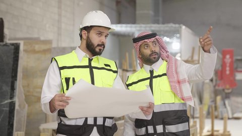 متابعة الأداء والمهام، حوارات ونقاشات هادفة، تصويرمقرب لمهندس عربي خليجي سعودي يرتدي سترة وخوذة الحماية يحمل مخطط هندسي برفقة زميله، الاشراف على سير خطة العمل، معامل ومصانع محليه سعوديه،