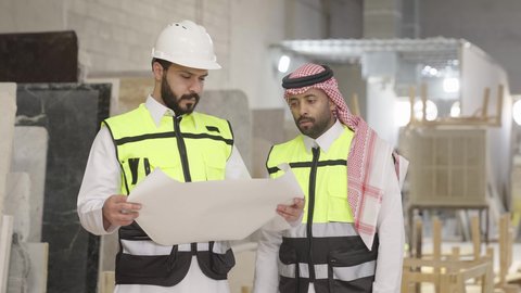 متابعة تنفيذ الخطط والمشاريع ، حوارات ونقاشات هادفة، تصويرمقرب لمهندس عربي خليجي سعودي يرتدي سترة وخوذة الحماية يحمل مخطط هندسي برفقة زميله، الاشراف على سير خطة العمل، معامل ومصانع محليه سعوديه،