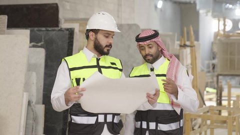 إدارة سير المشاريع والإشراف على التفاصيل، تصويرمقرب لمهندس عربي خليجي سعودي يرتدي سترة وخوذة الحماية يحمل مخطط هندسي برفقة زميله، الاشراف على سير خطة العمل، معامل ومصانع  محليه سعوديه، حوارات ونقاشات هادفة