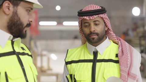 الإشراف والنقاش في العمل، تصوير مقرب لمهندسان سعوديان عاملان يشرفان في داخل المصنع في المملكة العربية السعودية , إرتداء خوذة وسترة الحماية الخاصة بالعمل