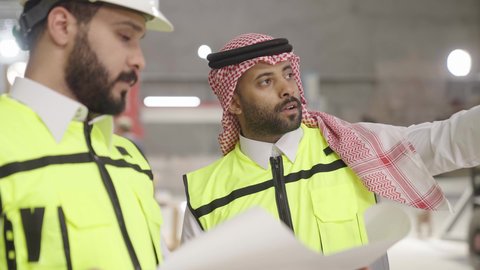Exchange of views, close-up shot of a Saudi Gulf Arab engineer wearing a vest and safety helmet holding an engineering plan alongside a colleague, supervising the progress of the work plan, local Saudi factories and plants, overseeing daily industrial operations.