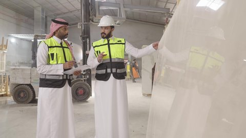 متابعة الأداء والإنتاج ، ارتداء خوذة الحمايه ، تصوير لمهندسان عربيان سعوديان خليجيان يرتديان الثوب يقومان بمناقشة العمل في احدى المستودعات ، معامل ومصانع محليه سعوديه، تسجيل و تدوين الملاحظات