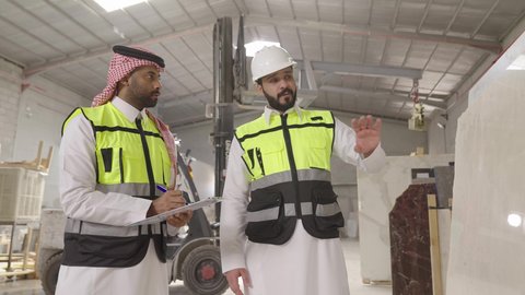 الإشراف على العمليات الصناعية اليومية، ارتداء خوذة الحمايه ، تصوير لمهندسان عربيان سعوديان خليجيان يرتديان الثوب يقومان بمناقشة العمل في احدى المستودعات ، معامل ومصانع محليه سعوديه، تسجيل و تدوين الملاحظات