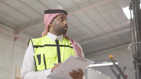 متابعة سير المشاريع، مهن ووظائف هندسية في المملكه، ايماءات التركيز في كتابة شيء ما، تصوير مقرب لمهندس عربي خيجي سعودي يرتدي ستره الحماية والشماغ يقوم بتدوين ونسجيل الملاحظات