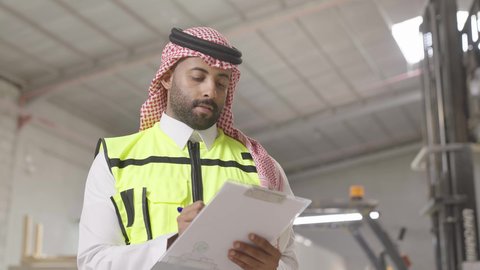 متابعة الأداء والإنتاج داخل المصنع، مهن ووظائف هندسية في المملكه، ايماءات التركيز في كتابة شيء ما، تصوير مقرب لمهندس عربي خيجي سعودي يرتدي ستره الحماية والشماغ يقوم بتدوين ونسجيل الملاحظات