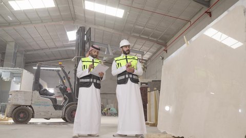 إدارة ومتابعة العمليات التشغيلية بالمصنع، ارتداء خوذة الحمايه ، تصوير لمهندسان عربيان سعوديان خليجيان يرتديان الثوب يقومان بمناقشة العمل في احدى المستودعات ، معامل ومصانع  محليه سعوديه، تسجيل و تدوين الملاحظات