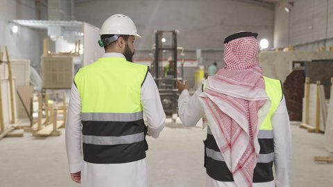 الإشراف على سير العمل في المنشأة الصناعية، ارتداء خوذة الحمايه ، تصوير من الخلف لمهندسان عربيان سعوديان خليجيان يرتديان الثوب يقومان بالمشي في احدى المستودعات ، معامل ومصانع  محليه سعوديه