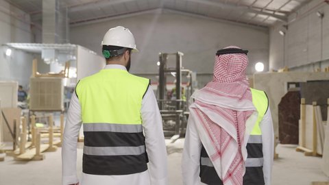 Monitoring the production steps in the factory, wearing safety helmets, a rear shot of two Arab Saudi Gulf engineers wearing traditional attire walking in one of the warehouses, local Saudi factories and plants.