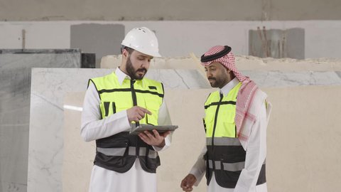 Modern technological devices, facilitating the performance of daily work tasks via tablets, youth professions and jobs, a Saudi Gulf Arab engineer wearing a safety vest and helmet is monitoring the progress of the factory's work alongside his colleague.