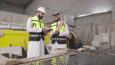 تسهيل متابعة وإنجاز الأعمال اليومية عبرالجهاز اللوحي، استخدام الأجهزة التقنية الحديثة، مهن ووظائف شبابيه، مهندس عربي خليجي سعودي يرتدي ستره الحماية و الخوذة يقوم بمتابعة سير أعمال المصنع برفقة زميله