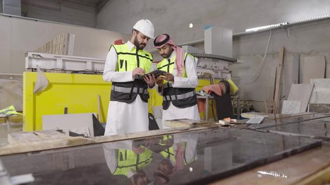 تبسيط المهام العملية اليومية باستخدام الجهاز اللوحي، استخدام الأجهزة التقنية الحديثة، مهن ووظائف شبابيه، مهندس عربي خليجي سعودي يرتدي ستره الحماية و الخوذة يقوم بمتابعة سير أعمال المصنع برفقة زميله