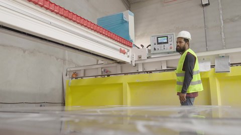 Saudi engineering professions and jobs, management and supervision of the manufacturing process, quality of industry and production in factories, productive Arab working hands, a Gulf Arab Saudi worker wearing a safety vest and a hard hat standing next to large machines.