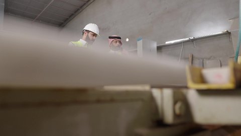 Easily completing daily tasks via tablet, using modern technological devices, youth professions and jobs, a Saudi Gulf Arab engineer wearing a safety vest and helmet is monitoring the progress of the factory's work alongside his colleague.
