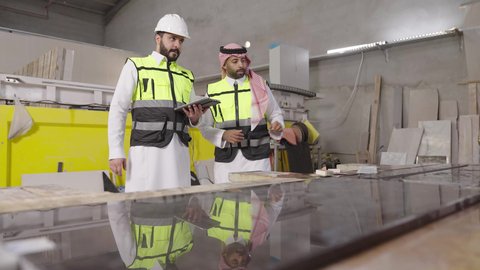 تسهيل القيام بمهام العمل اليومية عبرالتابلت، مهن ووظائف شبابيه، مهندس عربي خليجي سعودي يرتدي ستره الحماية و الخوذة يقوم بمتابعة سير أعمال المصنع برفقة زميله، استخدام الأجهزة التقنية الحديثة