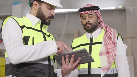 Youth professions and jobs, facilitating the daily work tasks via tablet, a close-up shot of a Saudi Gulf Arab engineer wearing a safety vest and helmet monitoring the progress of factory operations alongside a colleague, using modern technological devices.