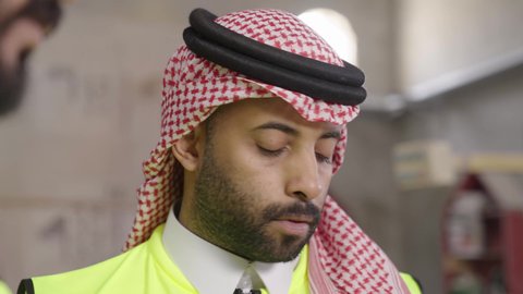 Organization and management of businesses, workshops, and factories in the Kingdom, close-up shot of a Saudi Gulf Arab engineer wearing a safety vest and a ghutrah supervising the progress of the work plan.