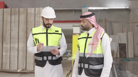 ورشات ومصانع في المملكة, تنظيم وإدارة الأعمال، تصوير لمهندس عربي خليجي سعودي يرتدي ستره الحماية و الخوذة يقوم تدوين وتسجيل الملاحظات على الحافظة برفقة زميله، الاشراف على سير خطة العمل