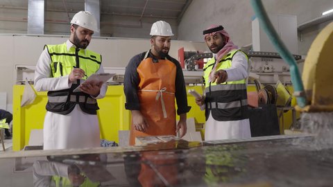 Achieving the industrial revolution, collaboration in task performance, a group of Arab Saudi Gulf industrial engineers wearing work clothes and safety helmets are monitoring the progress of the factory, the quality of production and manufacturing, Saudi professions and jobs, recording and documenting notes and information.