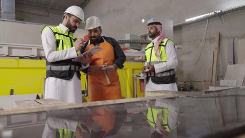 التعاون في أداء المهام، متابعه سير أعمال المصنع، مفهوم الاشراف على الانتاجية، مهندس عربي خليجي سعودي يرتدي سترة وخوذة الحماية يحمل بيده حافظة ورقية يقف برفقة عامل، جودة الانتاج والتصنيع، تحقيق الثورة الصناعية، مهن ووظائف سعودية