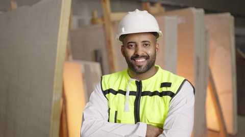 مهن ووظائف سعودية، تصوير لمهندس عربي خليجي سعودي مبتسم يرتدي الخوذة وسترة الحماية يقف بأيدي متقاطعه في موقع العمل ينظر إلى الكاميرا بإيماءات السرور، العمل في القطاع الهندسي