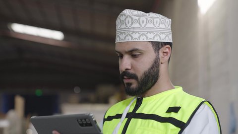 Operating the production line using a tablet device, modern technological and technical transformation, reliance on machinery and artificial intelligence in engineering, a close-up shot of an Arab Gulf Omani engineer wearing a safety vest and helmet monitoring the workflow inside the factory.
